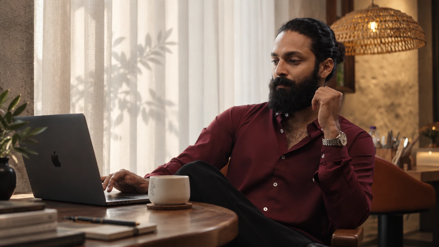 A man in a maroon shirt working on a laptop at a wooden desk